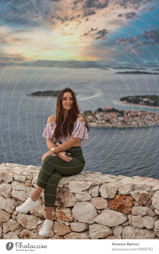 Attraktive Brünette, die an einem warmen, sommerlich windigen Tag auf einer Steinmauer sitzt. Modisches Mädchen posiert mit dem adriatischen Meer und der kleinen Stadt Primosten im Hintergrund. Wunderschöner goldener Sonnenuntergang