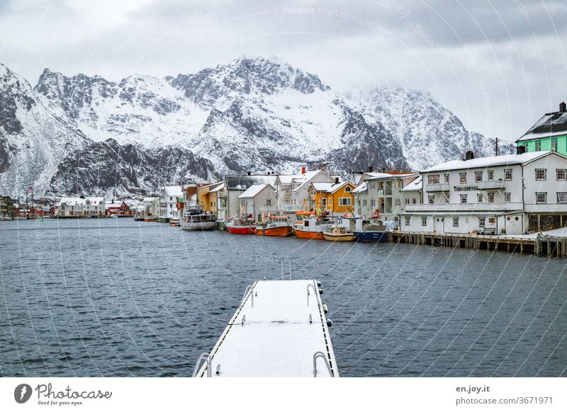 nordische Kleinstadt am Fjord mit Fischerbooten und Bootssteg vor verschneiten Bergen Henningsvær Lofoten Norwegen Fischerdorf Steg Bootsteg Schnee Winter Boote