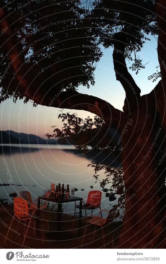 verlassener Tisch mit Flaschen, Gläser und Stühlen neben einem Baum an einem stillen See in Abenddämmerung Feierabend Abendlicht Sonnenuntergangsstimmung