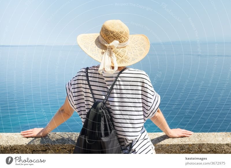 Rückansicht einer Reisenden mit Strohhut und Rucksack, die an einer Steinmauer lehnt und auf das große blaue Meer und die Inseln am Horizont schaut. Raum kopieren.