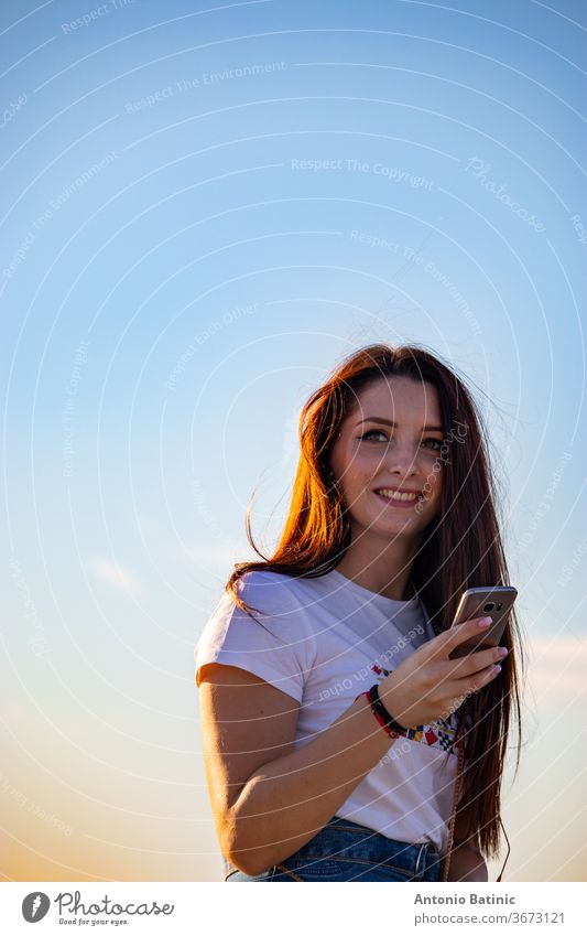 Attraktive Brünette, die ein Smartphone in der Hand hält und lacht, schöner sonniger Tag. Sonnenuntergang goldene Stunde, Haare leuchten orange von der Sonne, sauberer blauer Himmel