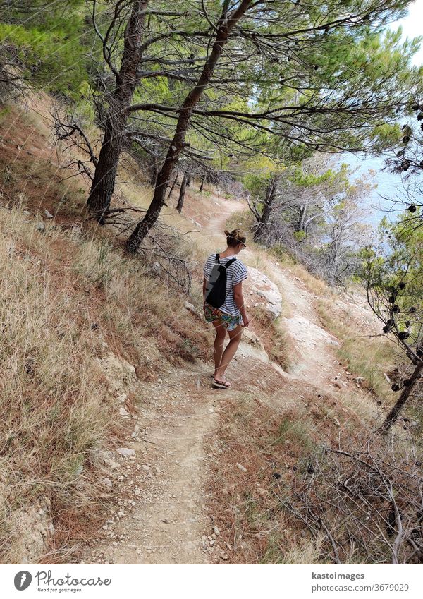 Junger aktiver Feamle-Tourist mit kleinem Rucksack, der auf einem Küstenweg zwischen Pinienbäumen wandert und nach einer abgelegenen Bucht sucht, um allein in Ruhe am Meer in Kroatien zu schwimmen. Reise- und Abenteuerkonzept.