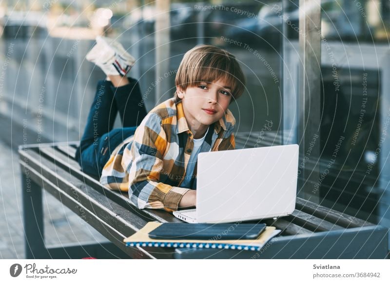 Ein lächelnder Junge liegt auf einer Bank und bereitet sich neben einem Rucksack auf den Schulunterricht vor. Der Junge ruht sich zwischen den Unterrichtsstunden aus. Soziale Distanz. Fernunterricht, Bildung, moderne Technologien