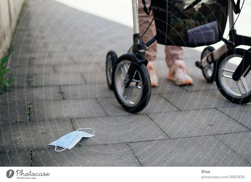 Eine Frau mit Behinderung geht mit ihrem Rollator, daneben ein am Boden liegender Mundschutz. Maskenpflicht um Risikopatienten zu schützen. Risikopatientin