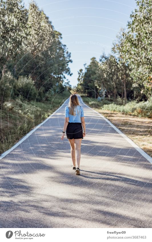 Junge kaukasische Frau geht an einer leeren Straße in der Nähe eines Waldes laufen Lifestyle Textfreiraum Landschaft Mode Model jung blond Kaukasier Sonnenlicht