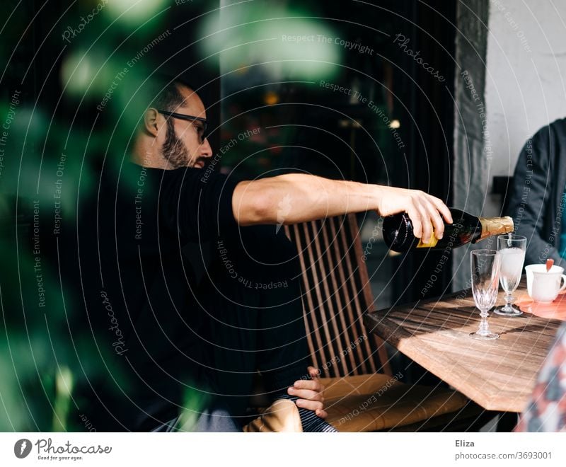 Ein Mann sitzt am Tisch im Garten und schenkt Prosecco in Gläser ein einschenken ausschenken Flasche Sekt Sektflasche Feier Daydrinking eingießen gemütlich