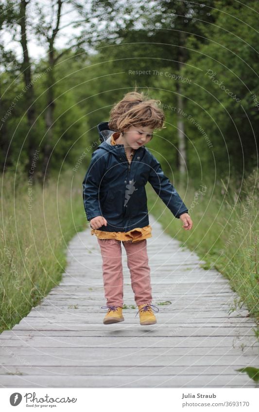 Kleinkind fliegend in der Luft in Regenjacke auf einem Holzbretterweg Mädchen frei Freude Haare wild Abheben lustig grinsen toben Spielen Wege & Pfade Holzsteg