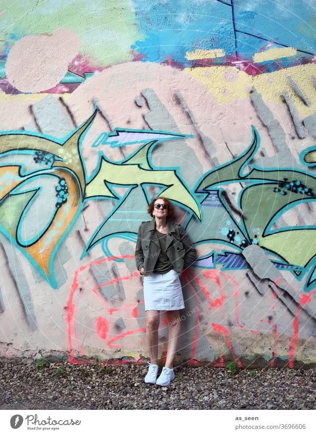 Frau mit Sonnenbrille vor einer Wand mit Graffiti Sneaker gelockt rothaarig Außenaufnahme Tag Mensch Mode grün khakigrün bunt Muster anlehnen cool Junge Frau