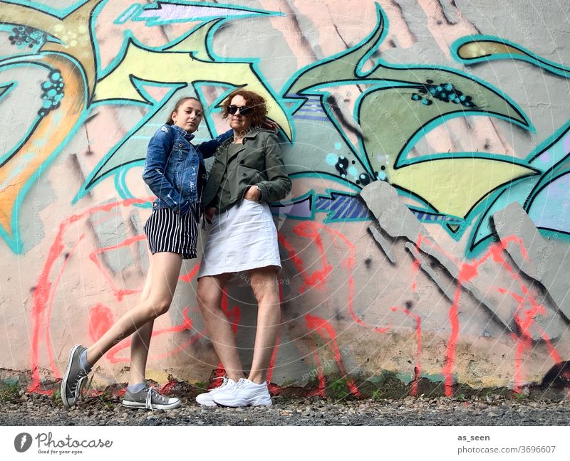 Zwei Frauen vor einer Wand mit Graffiti Sneaker Sonnenbrille gelockt rothaarig Außenaufnahme Tag Mensch Mode grün khakigrün bunt Muster anlehnen cool Coolness