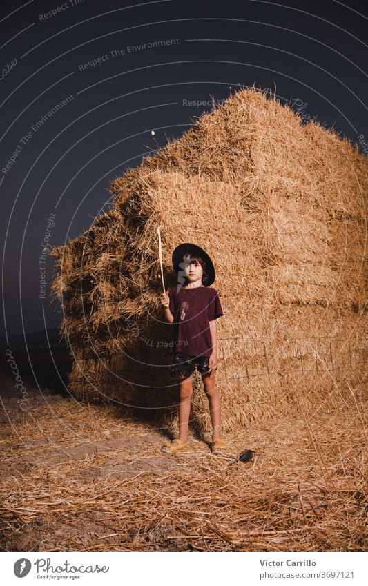 Ein Junge mit Hut, der im Sommer auf den Feldern auf dem Land in einem Strohballenhintergrund spielt Bauernbursche hockend Pflanze Spielen Herbst Kind niedlich