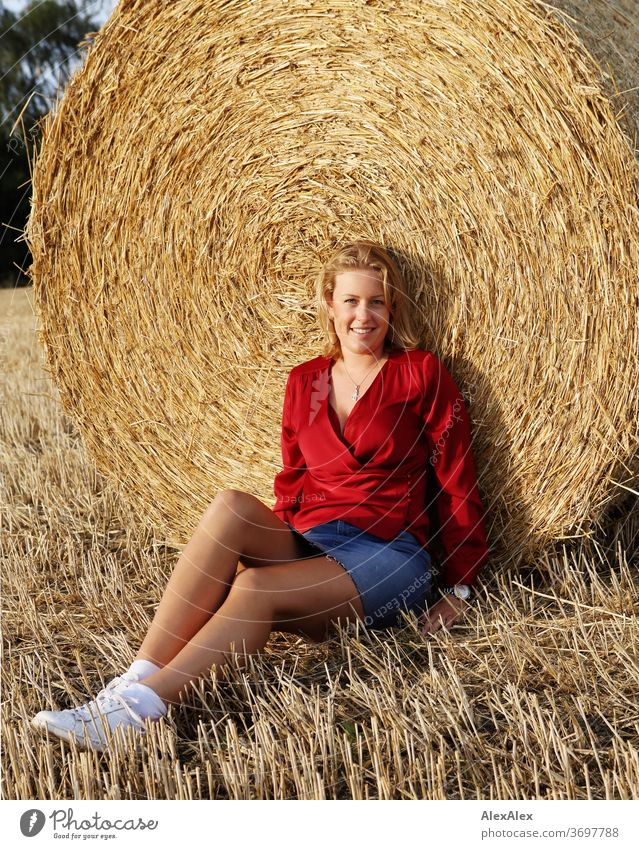 Portrait einer jungen Frau an einem Strohballen im Abendlicht junge Frau blond Lächeln rot Schmuck schön langhaarig Landschaft Wolken Himmel gebräunt