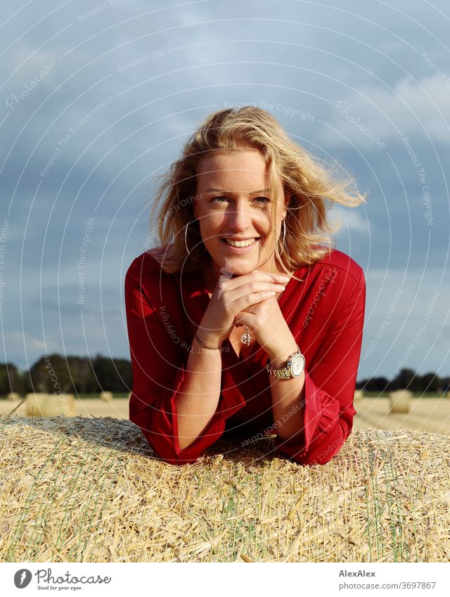 Portrait einer jungen Frau auf einem Strohballen im Abendlicht junge Frau blond Sommersprossen Lächeln rot Schmuck schön langhaarig Oberkörper Landschaft Wolken