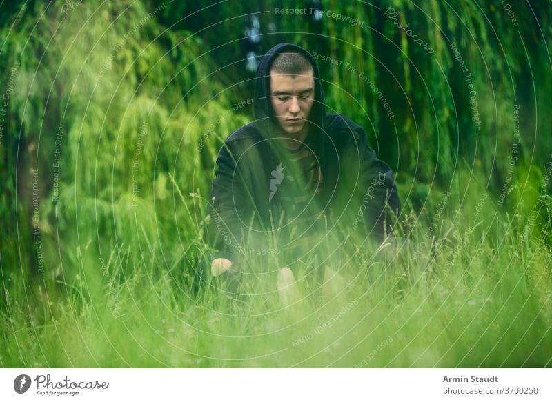 Porträt eines traurigen, kauernden Mannes in einem schwarzen Kapuzenpulli jung ernst Sorge hockend Wiese Natur Sommer Aussehen Teenager Blick männlich schön