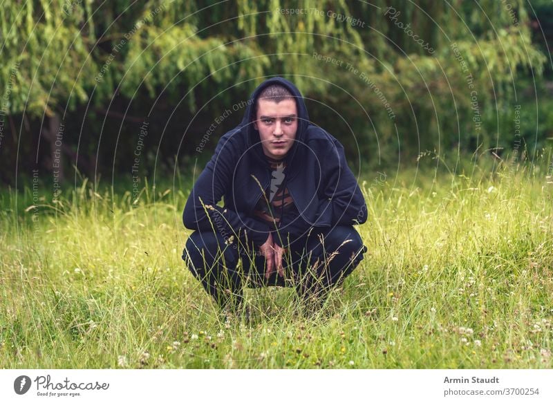 Porträt eines lächelnden, kauernden Mannes in einem schwarzen Kapuzenpulli jung Lächeln Glück glücklich hockend Wiese Natur Sommer Aussehen Teenager Blick