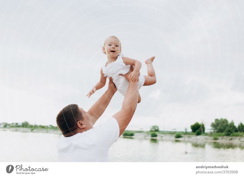 Vater wirft Baby auf Pier auf Seehintergrund, Mädchen fliegt in den Himmel. Portrait Vater mit Kind zusammen. Papa, kleine Tochter im Freien. Angelurlaub der Familie am Teich.