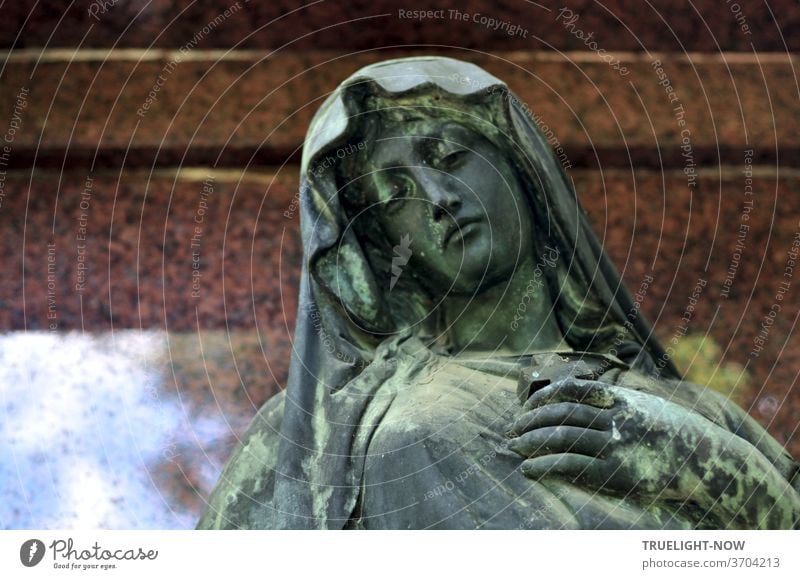 Blaugrün schimmernde Skulptur einer schönen trauernden Frau vielleicht aus der Jugendstil Epoche vor rötlich braun poliertem Marmor, in dem sich ein wenig blauer Himmel mit weissen Wolken spiegelt, was der Trauer aber nicht schadet