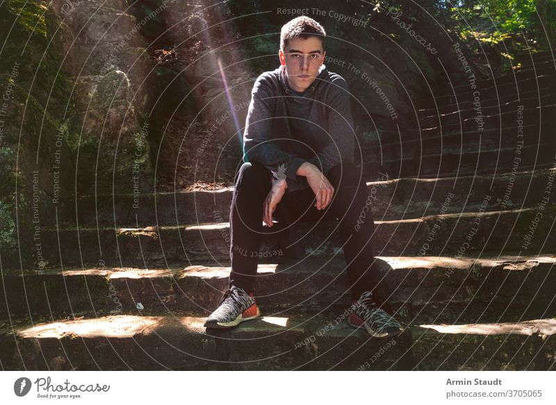 Porträt eines ernsthaften jungen Mannes, der auf einer Treppe sitzt Sitzen bw cool lässig Sonnenlicht Sonnenstrahlen im Freien Perspektive trendy selbstbewusst