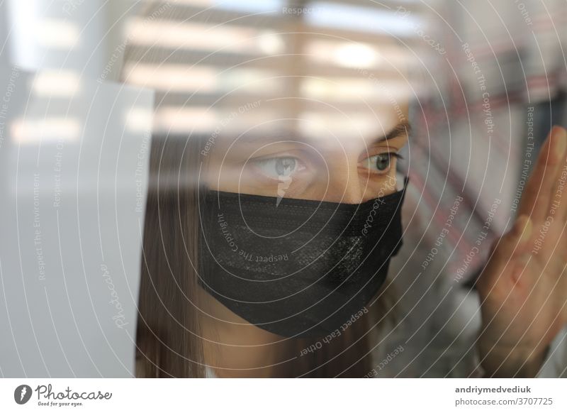 junge frau in einer medizinischen maske hinter einer fensterglasscheibe zu hause. konzept der quarantäne der coronavirus-pandemie, covid 19. selektiver fokus