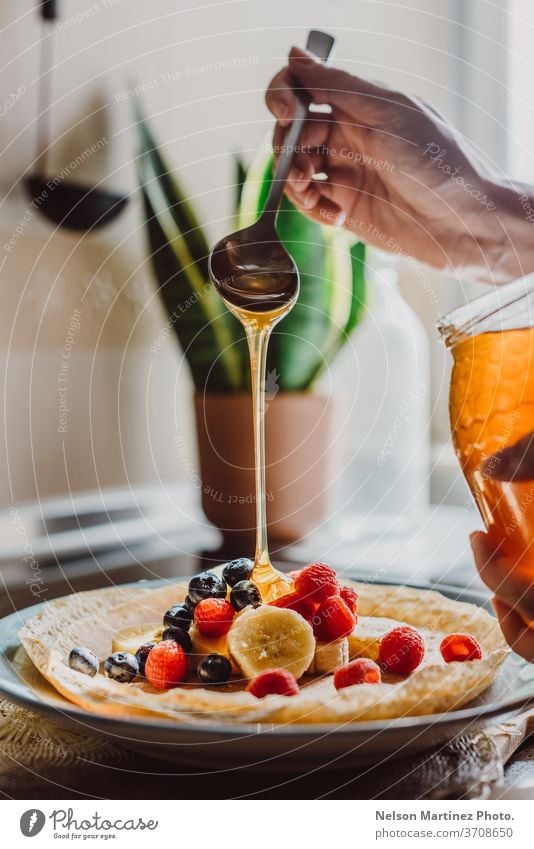 Köstlicher Crêpe mit Bananen, Heidelbeeren, Himbeeren und Honig. Die Küche ist mit einem schönen natürlichen Licht beleuchtet. Wir können sehen, wie der Honig vom Löffel fällt.