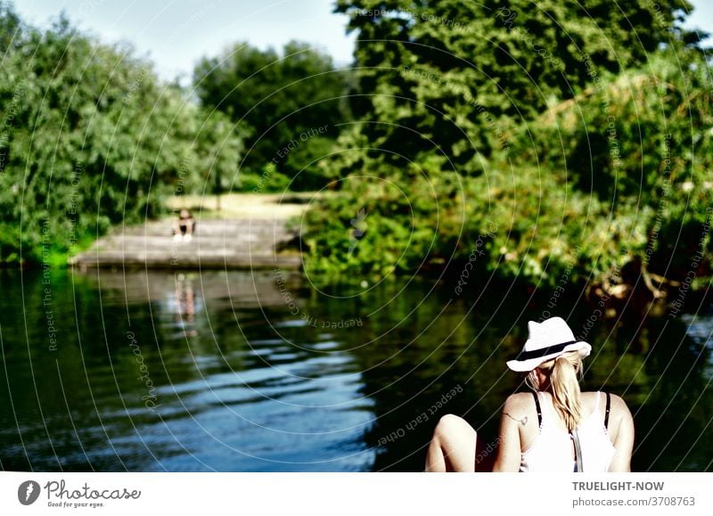 Die Sonne scheint auf die helle Haut nackter Schultern und des nackten, angewinkelten Beins einer jungen Frau mit Strohhut und blondem Pferdeschwanz, die auf der Kaimauer sitzend ins Wasser vor dem Ufer der baumbestandenen Freundschaftsinsel schaut