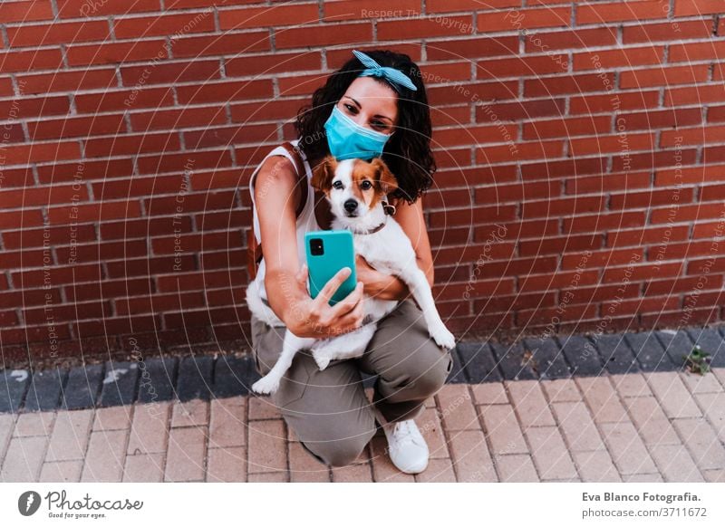 junge Frau, die mit einer Schutzmaske im Freien spazieren geht und mit ihrem Handy ein Foto des süßen Hundes Jack Russell macht. Neues normales Konzept Straße