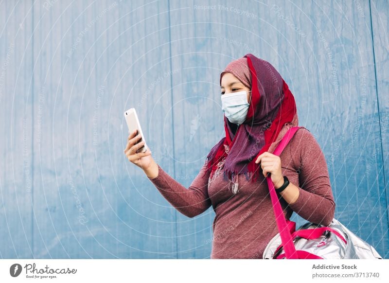 Sportliche Frau in Maske mit Smartphone auf der Straße Sportlerin Großstadt benutzend Hijab Mundschutz medizinisch Coronavirus Sportbekleidung arabisch ethnisch