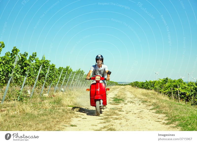 Roller im Weinberg fahren. Mann, mit Helm der Roller in der Landschaft fährt. Porträt eines Mannes und mit einem stilvollen Vintage-Moped im Sommer gegen blauen Himmel