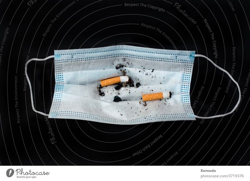 Gesichtsmaske mit Zigarettenstummeln als Aschenbecher, isoliert auf Schwarz. Mundschutz Coronavirus Bund 19 Korona Missbrauch neue Normale dumm Konzept Rauch