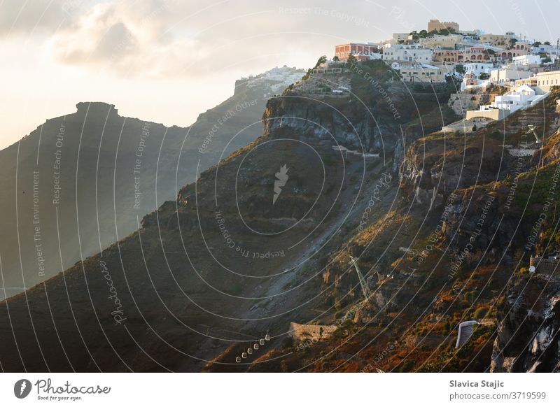 Sonnenuntergang auf der griechischen Insel Santorin mit farbenfrohem, warmem Licht und Wolken über der Stadt. Der Blick folgt dem Rand der Caldera von Thira, Skaros-Felsen und Imerovigli in Richtung Sonne