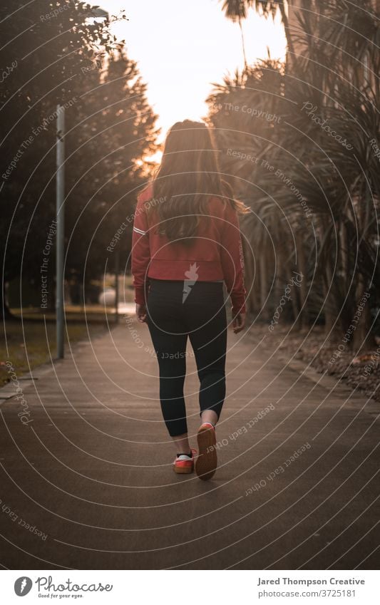 Eine junge Frau begibt sich auf einen von Bäumen gesäumten Weg, gebadet von der warmen Glut des Sonnenuntergangs. rot glühen Wärme lange Haare Mädchen Teenager