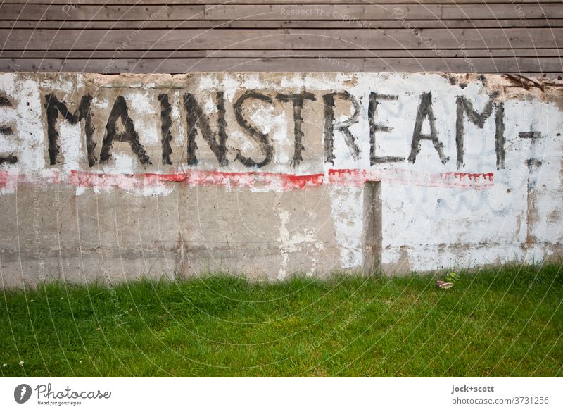 Mainstream auf Fundament Beton Wort Wiese Großbuchstabe Straßenkunst Subkultur Kreativität Holzzaun Anstrich lost places Wand Rest Pinselstrich verwittert