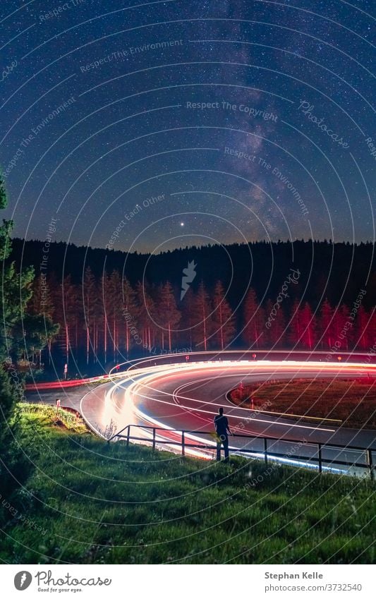 Milkyway, Langzeitbelichtung eines Mannes, der an einem Geländer vor einer kurvigen Straße steht, während Autos vorbeifahren und das Licht der Karrenbahn verlassen.