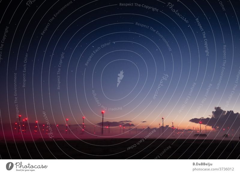 Mystische rote Punkte im Morgengrauen. Unter einem klaren Sternenhimmel schmiegt sich der aufsteigende Nebel an die beleuchtete Säulen des Windparks in Ostfriesland.