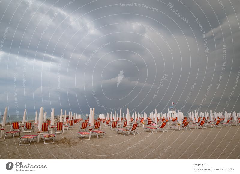 s'isch over - Sommerurlaub - Strand mit Liegestühlen und zugeklappten Sonnenschirmen, da ein unwetterartiges Gewitter mit dunklen Wolken bis zum Horizont am Himmel aufzieht