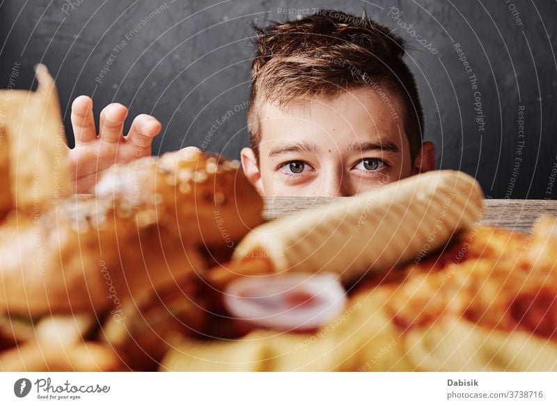 Ein hungriger Junge schaut sich leckeres Essen an und nimmt einen Hamburger vom Tisch. Konzept ungesundes Essen Fett Diät Burger Pizza Hintergrund Cholesterin