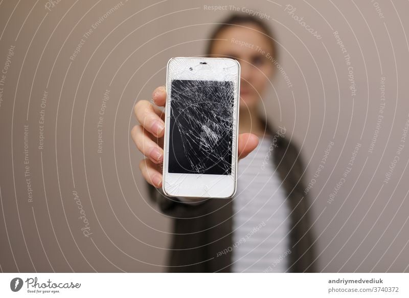 Frustrierte junge Frau hält Handy mit zerbrochenem Bildschirmglas in der Hand. Das Display des Telefons muss repariert werden. Isoliert auf grauem Hintergrund. selektiver Fokus auf Smartphone