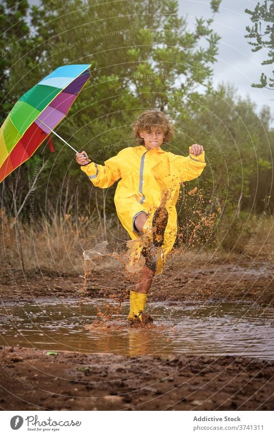 Junge mit Stiefeln und Regenmantel hält einen Regenschirm und spielt in einer Pfütze Witz Schuljunge Dusche Einstellung Regenbogen lockig gestikulieren