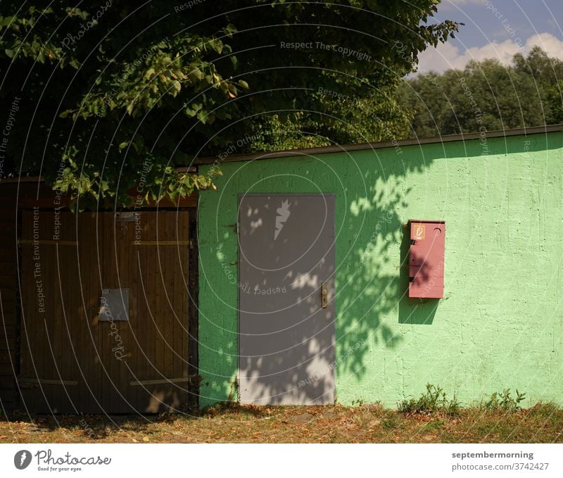 Garage mit Anbau altes Garagentor mit angeschlossenem seitlichen Gebäude. braue Tür grüne Fassade Feuerlöscher Baum Schattenbildung Sonnenlicht Wärme