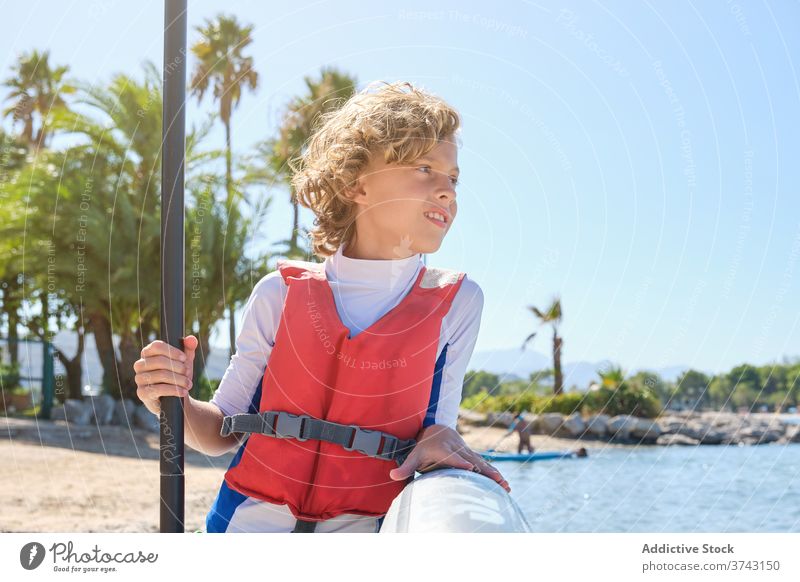 Abgelenkter Junge mit Weste, der sich an ein Paddel-Surfbrett lehnt und den Paddelstock hält retten Übung Kind Outfit Surfen Ruhe trainiert. behüten schützend