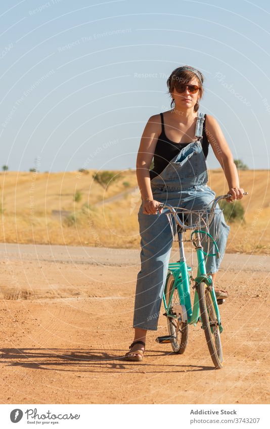 Ruhige Frau auf dem Fahrrad in ländlicher Umgebung Straße Sand Sonnenlicht gesamt Landschaft Gelassenheit Sonnenbrille Jeansstoff Sommer Natur