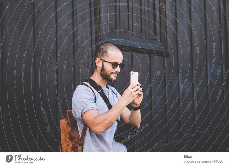 Ein Mann mit Sonnenbrille, Rucksack und professioneller Kamera fotografiert etwas mit einem Smartphone Tourist fotografieren Ausflug Reisender Fotograf lächeln