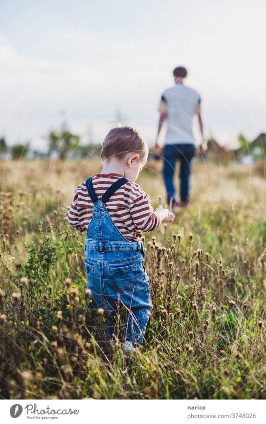 Kleiner Junge läuft über Wiese, Vater im Hintergrund Kleinkind Kind Außenaufnahme Kindheit Farbfoto 1-3 Jahre Natur Latzhose Leben Mensch Schwache Tiefenschärfe