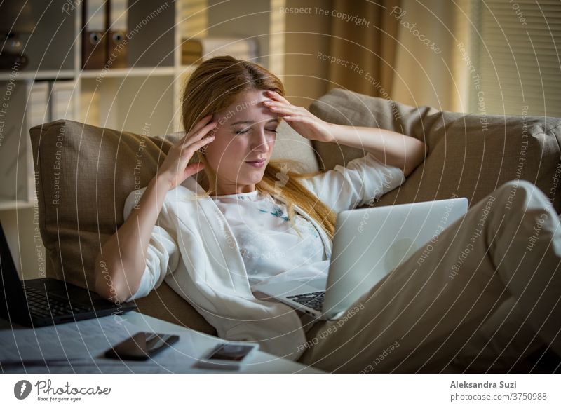 Attraktive Geschäftsfrau auf einer Couch mit zwei Mobiltelefonen und Laptops. Erschöpfte Frau im Büro spät in der Nacht. Verantwortliche Führungskraft bei der Arbeit, Kopfschmerzen, Hände an den Schläfen