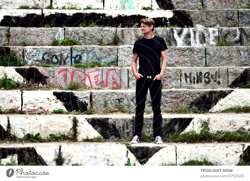 Tim Trzoska, Berlin Fotograf,  Berlin Kenner von Geburt, steht in schwarzen Jeans, schwarzem T-Shirt und leichten Sneakers auf mit Graffiti bemalten Stein-Stufen am Mauerpark in Berlin Prenzlauer Berg