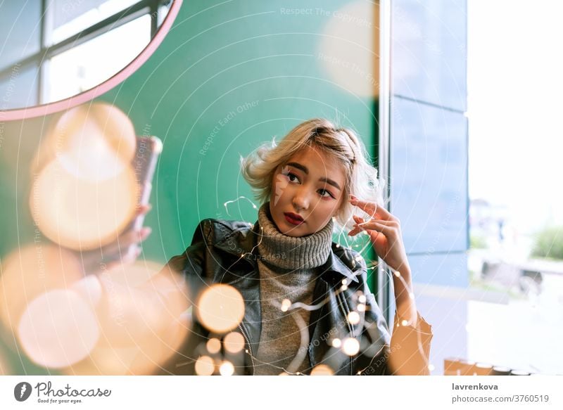 Junge Asiatin mit blonden Haaren beim Selfie mit Lichterkette Winter Herbst Weihnachten saisonbedingt Erwachsener asiatisch Bokeh Café Weihnachtsbeleuchtung