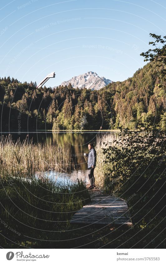 Person trinkt Kaffee am See vor Bergkulisse Allgäu Oberstdorf Gipfel Skischanze Wald Wasser Steg Frau jugendlich Kaffeetasse Camper Outdoor Style Holzsteg