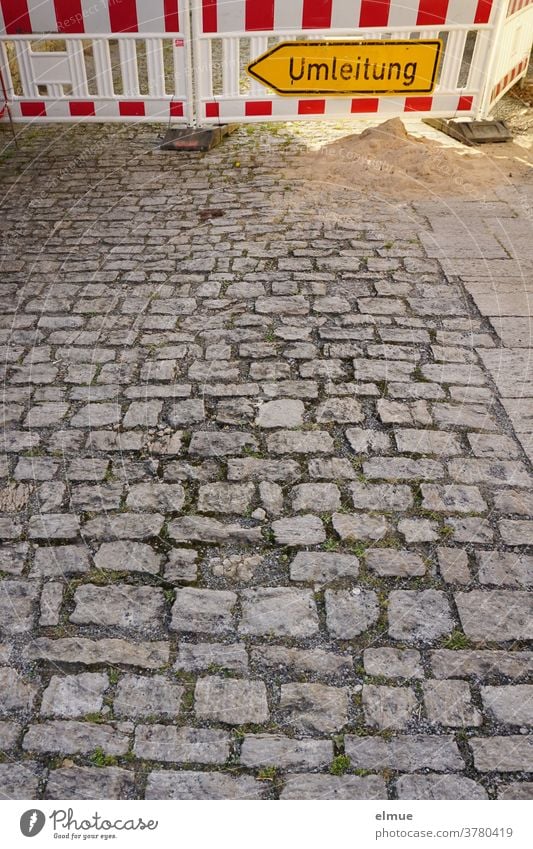 Ein gelbes Umleitungsschild und rot-weißes Absperrgitter vor einer Baustelle verhindern das Weiterfahren auf der grauen , gepflasterten Straße. Schild