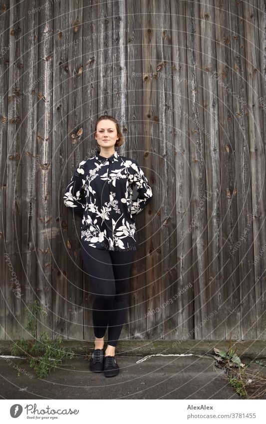 Junge Frau lehnt an einer Bretterwand und schaut in die Kamera Erwartung Farbfoto Blick in die Kamera einzigartig Sommer Außenaufnahme selbstbewußt sportlich