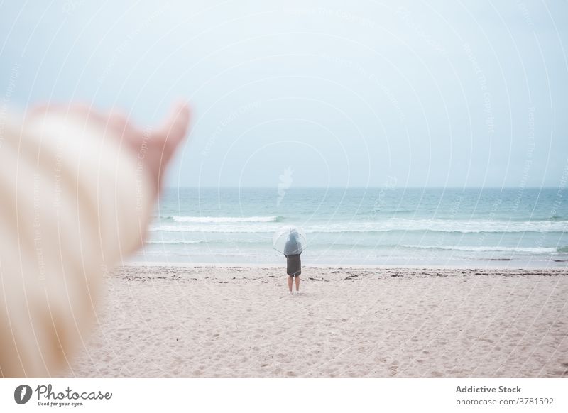 Unbekannter Reisender mit Regenschirm bewundert das Meer Strand MEER Zeigen Sie weg Sand reisen Ufer Küste bedeckt einsam Natur winken Wetter Tourismus Seeküste