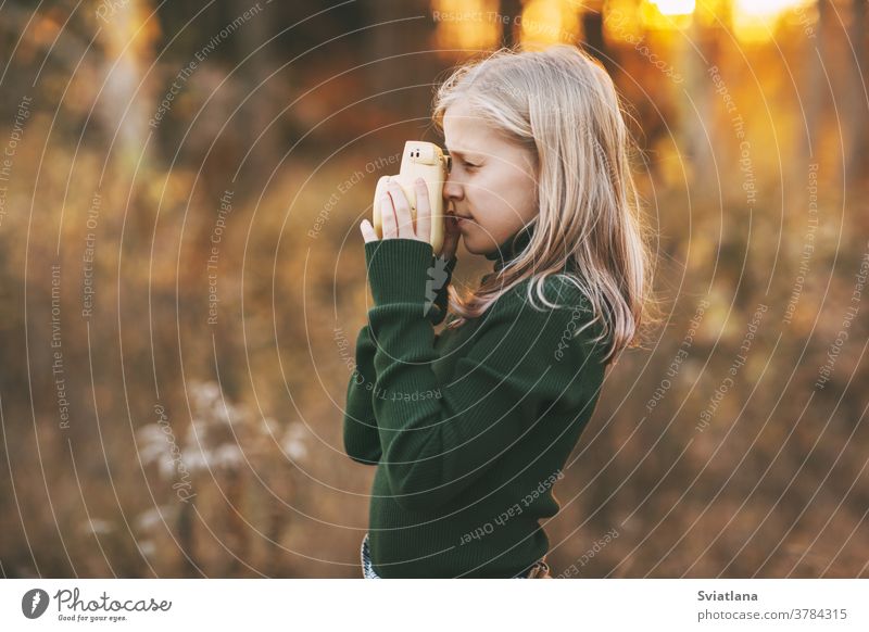 Ein hübsches Teenagermädchen mit blonden Haaren fotografiert die Natur mit einer gelben Polaroidkamera in einem Herbstpark. Seitenansicht. traurig besinnlich